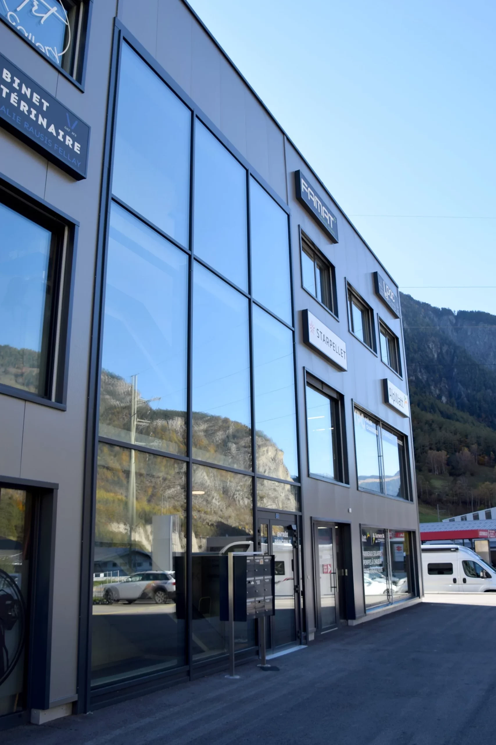 Grandes fenêtres vitrées sur une façade contemporaine grise, avec des montagnes en arrière-plan.