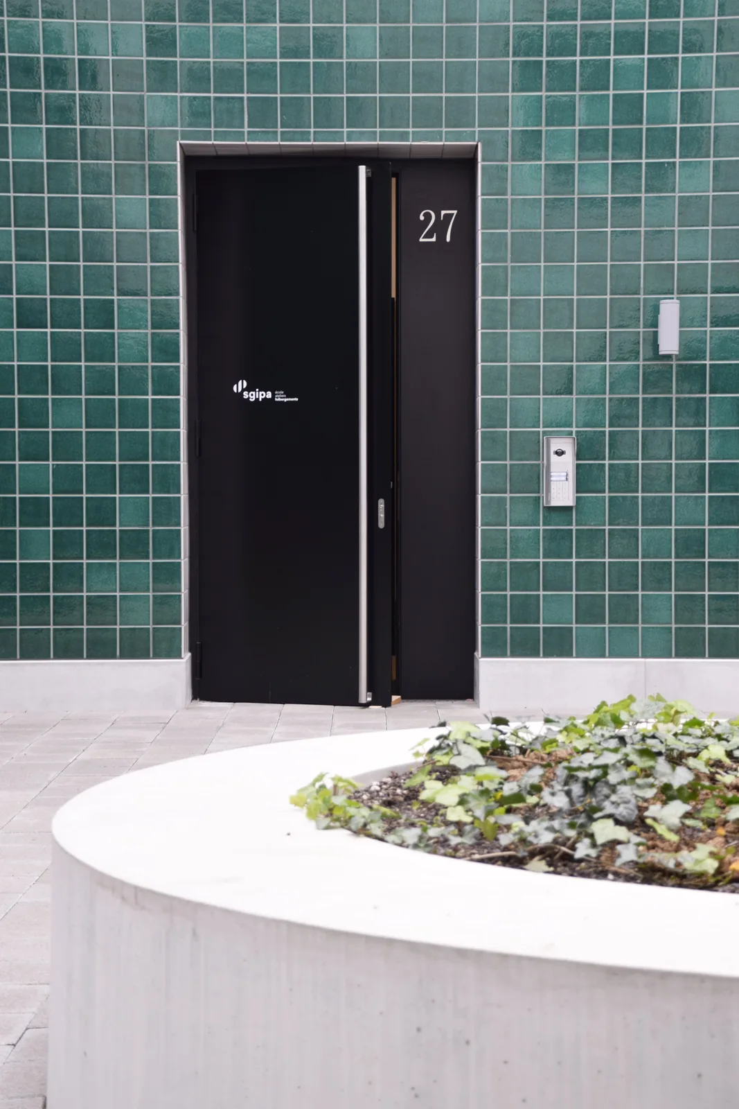 Porte d’entrée anthracite entourée de murs carrelés verts et de vitrages.